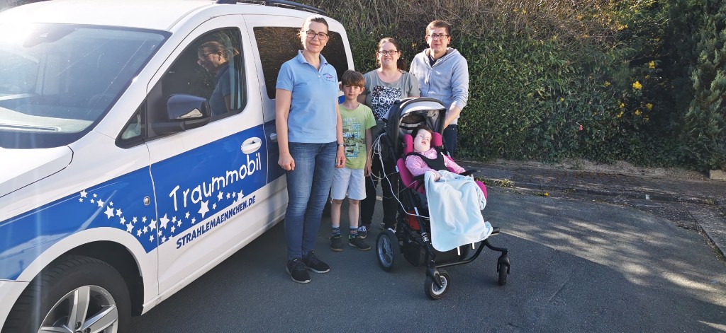 Familie steht gemeinsam mit dem Team am Traummobil, ein Kind sitzt im Spezial-Buggy