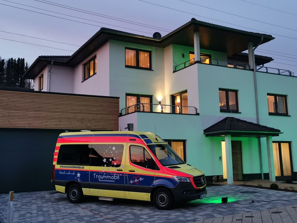 Haus Konrad am Abend: die grün angestrahlte Fassade mit erleuchteten Fenstern, davor das Traummobil in der Einfahrt