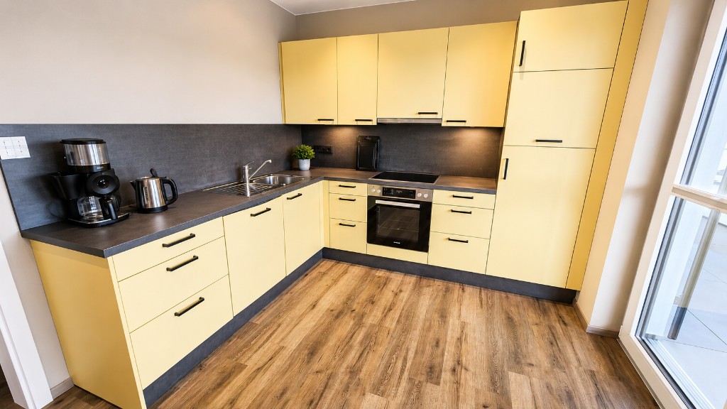 Küche einer Familienwohnung in Haus Konrad: hellgelbe Einbauküche über Eck mit Backofen, Ceranfeld, Dunstabzug, Spüle, hohem Kühlschrank, Kaffeemaschine und Wasserkocher, daneben ein bodentiefes Fenster