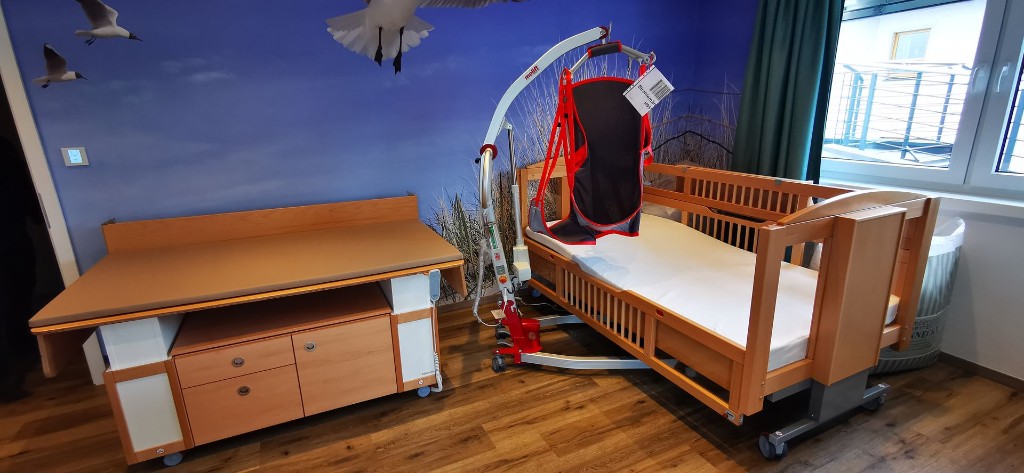 Pflegezimmer in Haus Konrad mit höhenverstellbarem Pflegebett, Patientenlifter, Pflegeliege und einem Wandbild mit Meer und Möwen