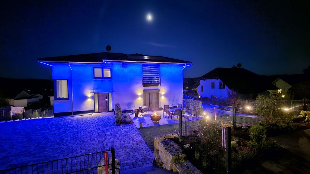 Gartenseite von Haus Konrad am Abend: blau angestrahlte Rückseite des Hauses, davor die Terrasse mit Sitzgruppe und Feuerschale und der eingezäunte Garten