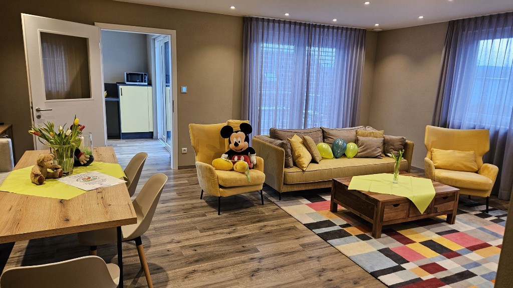 Wohnzimmer einer Familienwohnung in Haus Konrad mit Sofa, zwei Sesseln, bunt gemustertem Teppich, gedecktem Esstisch mit Blumen und offenem Durchgang zur Küche